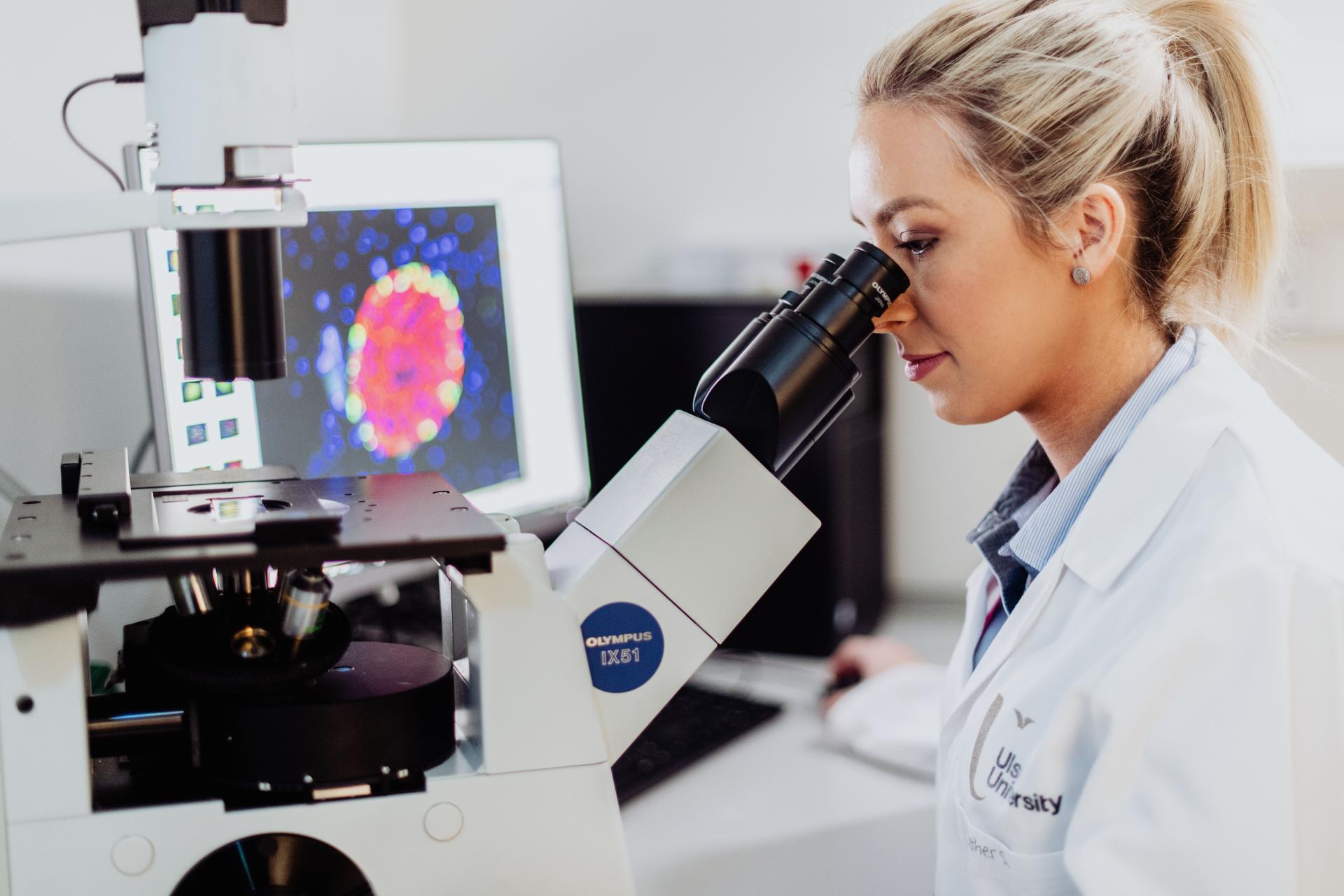 Person in a white Ulster University laboratory coat using a microscope in a biomedical research laboratory. A computer monitor in the background displays a colourful microscopic image, while laboratory equipment and workstations are visible in the bright, modern research setting.